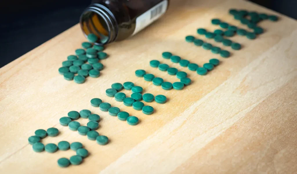 Tabletas verdes que deletrean 'Espirulina' sobre una superficie de madera con un frasco de medicina cerca, que representa suplementos nutricionales para atletas.